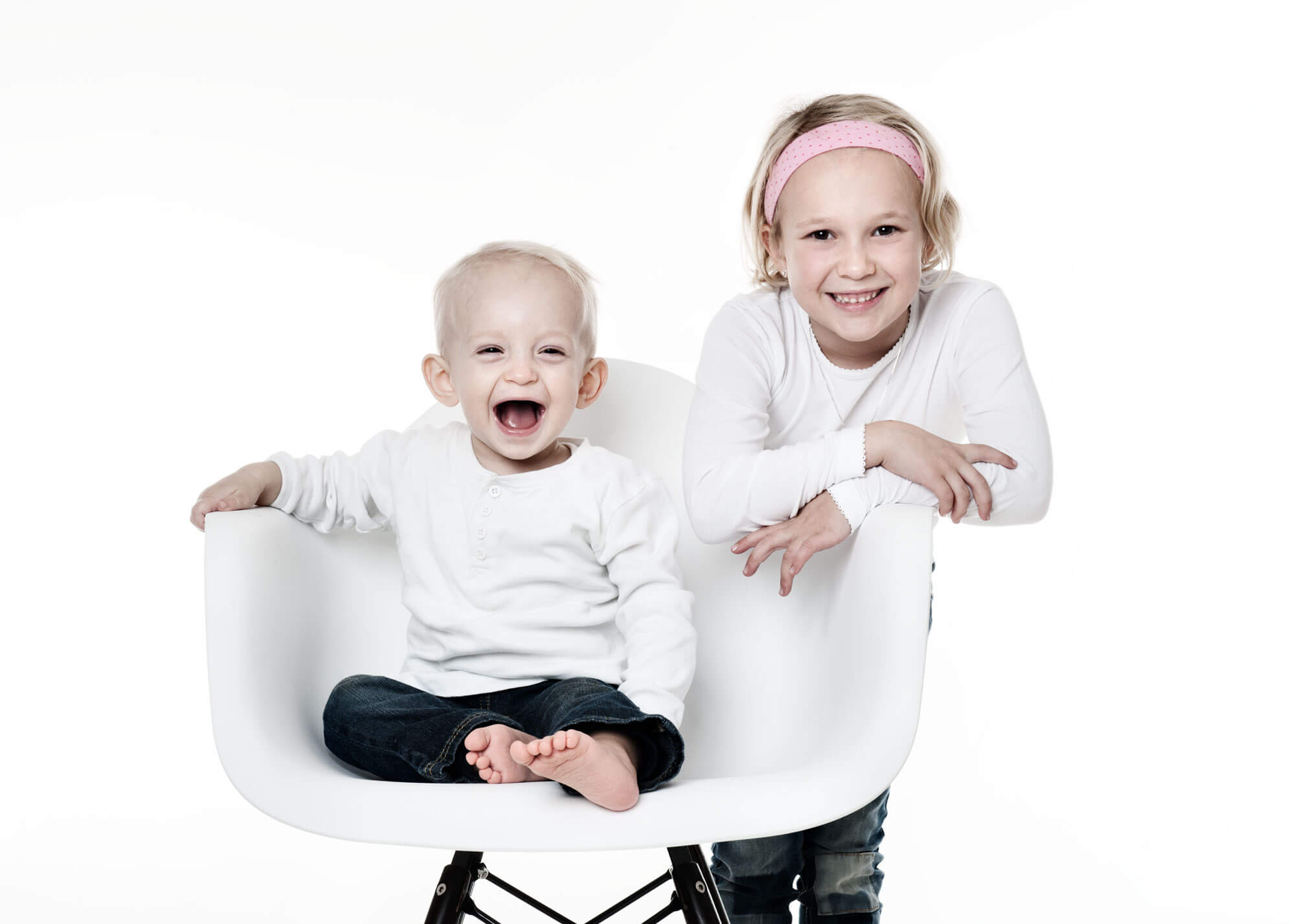Kleinkind sitzt herzlich lachend auf einem weißen Stuhl. Die große Schwester kuschelt sich von hinten lachend an